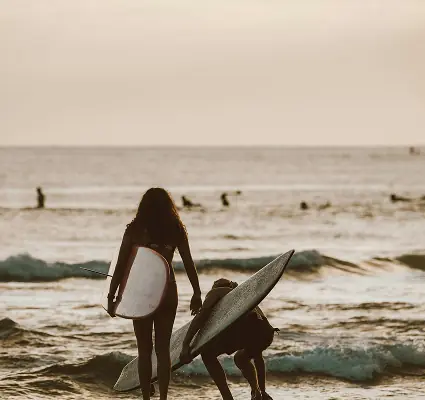 Two surfers entering the ocean at sunset with other surfers in the background