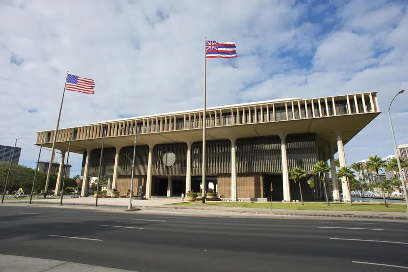 Hawaii State Capitol building with U.S. and Hawaii flags against a cloudy sky