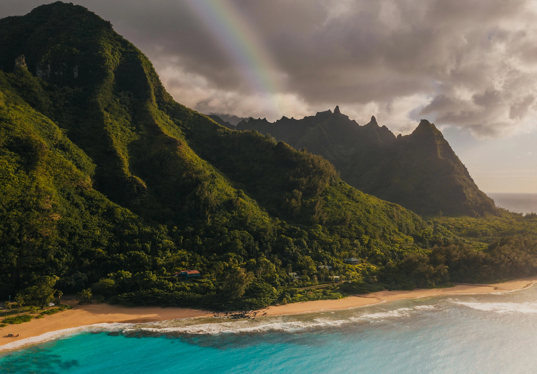 Green mountains and a sandy shoreline with a faint rainbow overhead