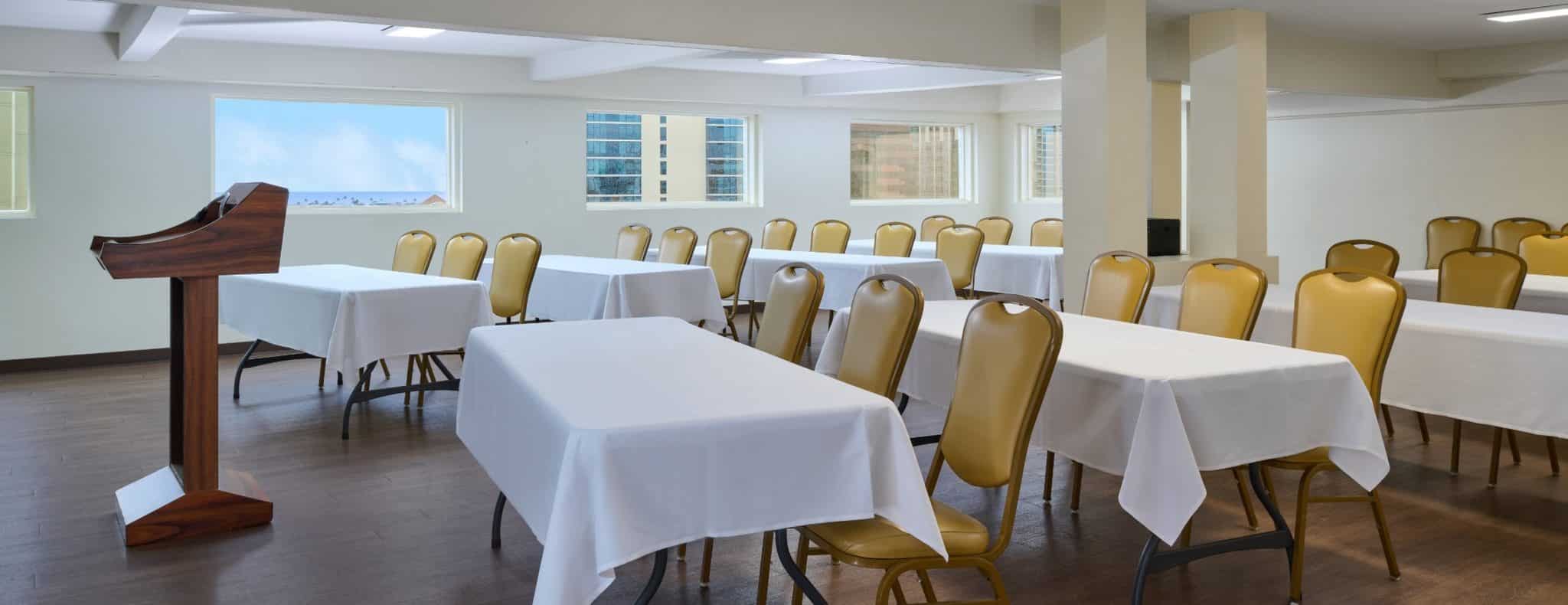 The Penthouse meeting room at Pagoda Hotel set with tables, gold chairs and a podium, with windows looking out to the ocean and Honolulu skyline